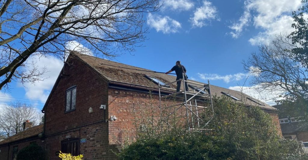 Scraping the moss off a slate roof to clean it and improve aesthetics. The worker is stood on a scaffold access tower for safety and using a special scraper blade and a telescopic pole